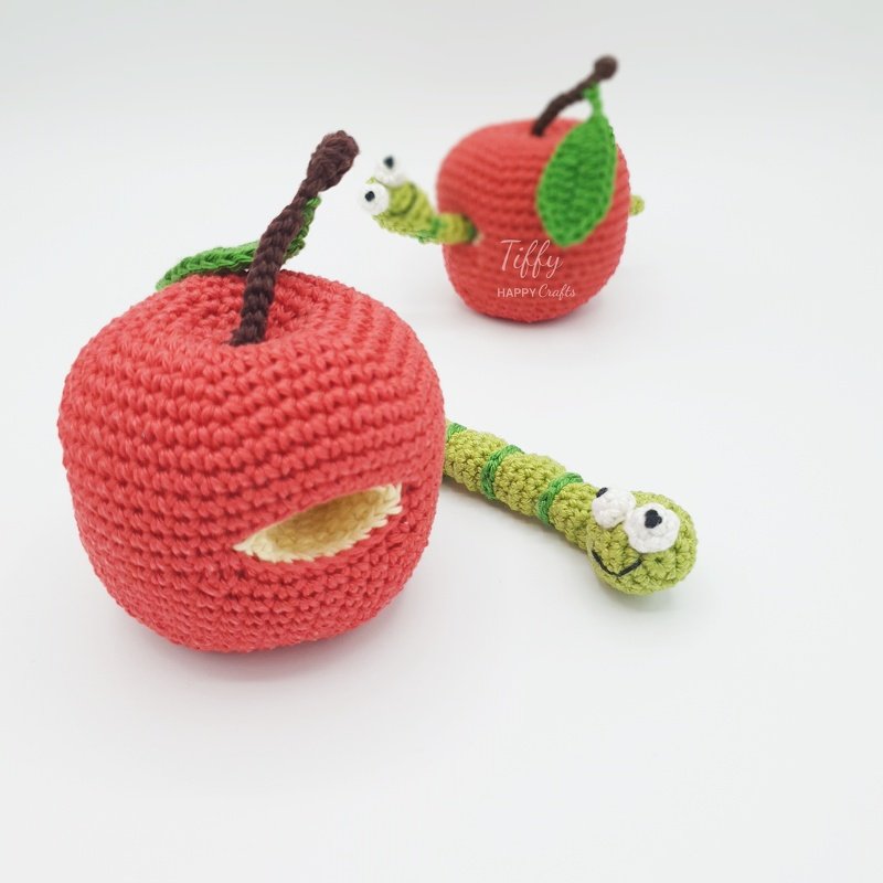 Two red crocheted amigurumi apples with green worms; one worm emerging from a small apple and a longer worm beside a larger apple with a hole.