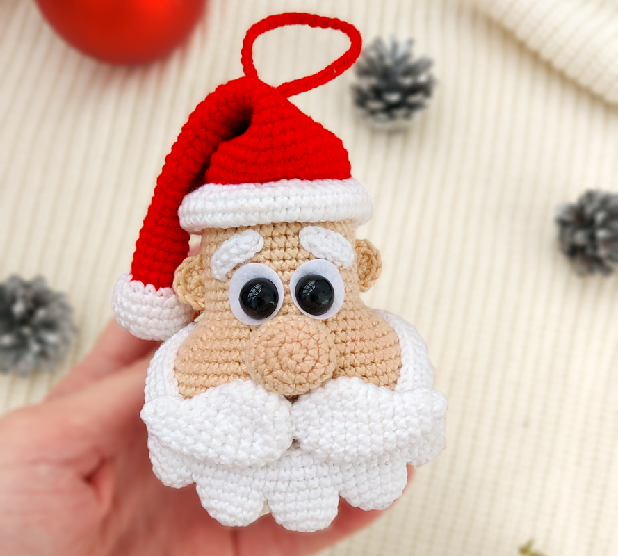 Hand holding a crocheted Santa Claus head ornament with red hat, white beard and googly eyes