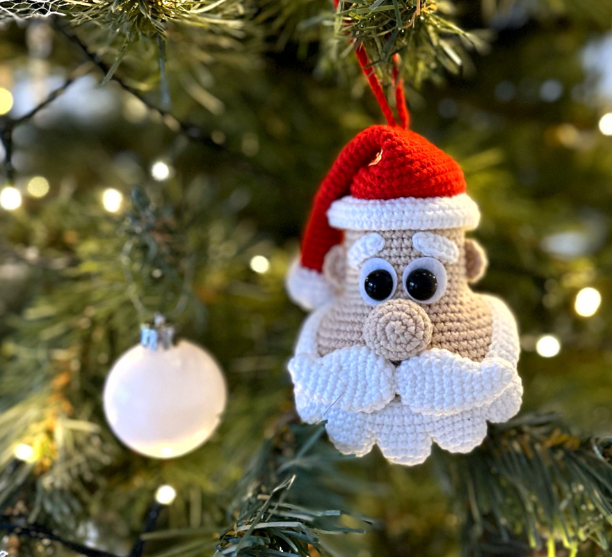 Crocheted Santa Claus ornament with red hat, white beard, and large black eyes hanging on a Christmas tree.
