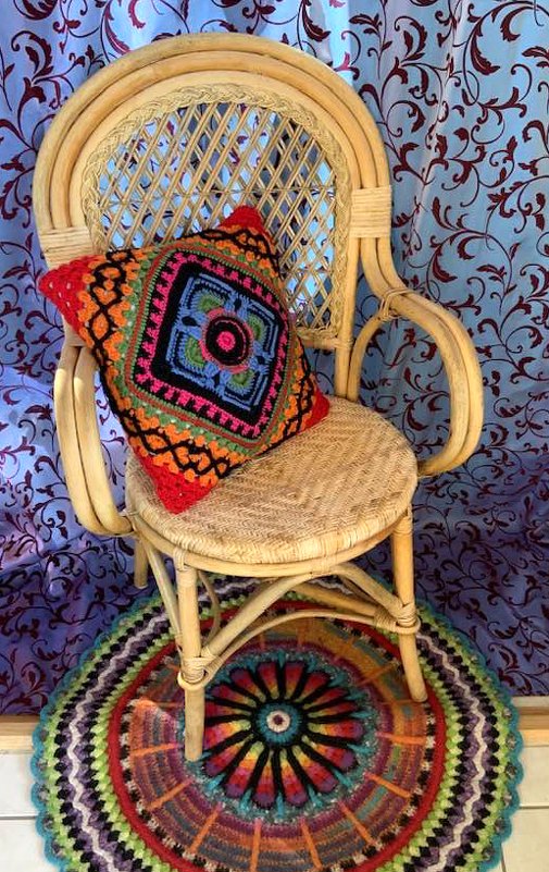Gehäkeltes buntes Kissen auf hellem Rattansessel, runder Häkelteppich und gemusterter Vorhang.