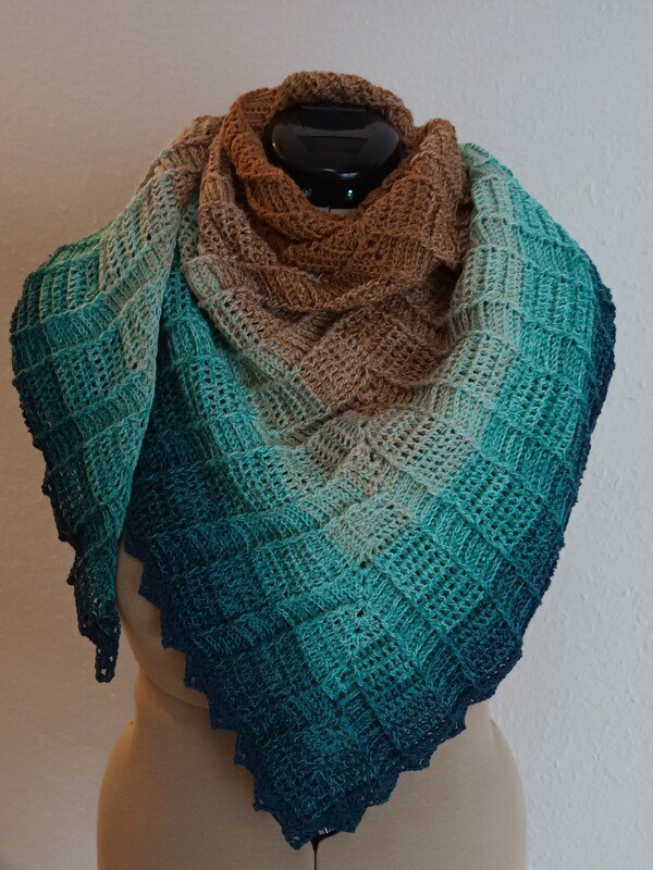 Gehäkeltes Dreieckstuch mit Farbverlauf von Braun zu Türkis und Blau, quadratisches Strukturmuster, auf einer Schneiderpuppe drapiert.