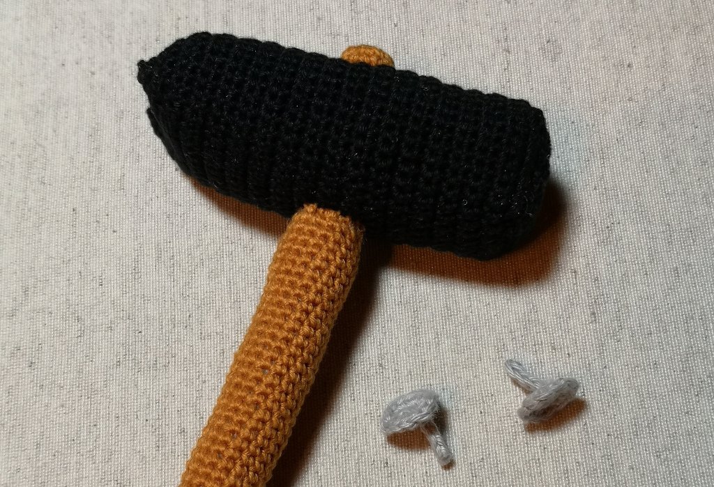 Gehäkelter Hammer mit schwarzem Kopf und braunem Stiel neben zwei kleinen gehäkelten Nägeln auf hellem Hintergrund.