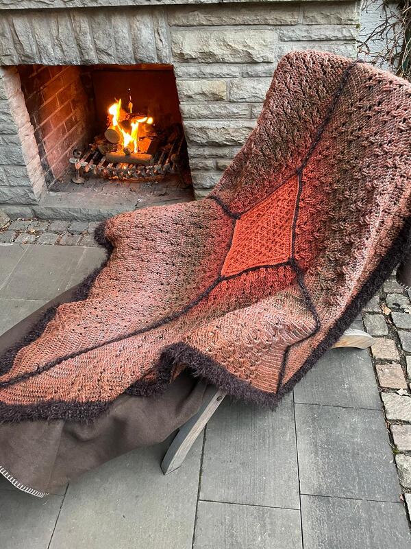Braun-orange gehäkelte Decke mit Rautenmuster auf einem Stuhl vor steinernem Kamin