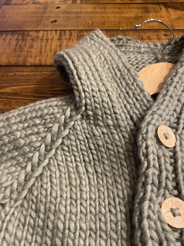 Detailansicht einer grauen Kinderstrickjacke mit Raglanärmelnaht, sichtbaren Maschen und Holzknöpfen.