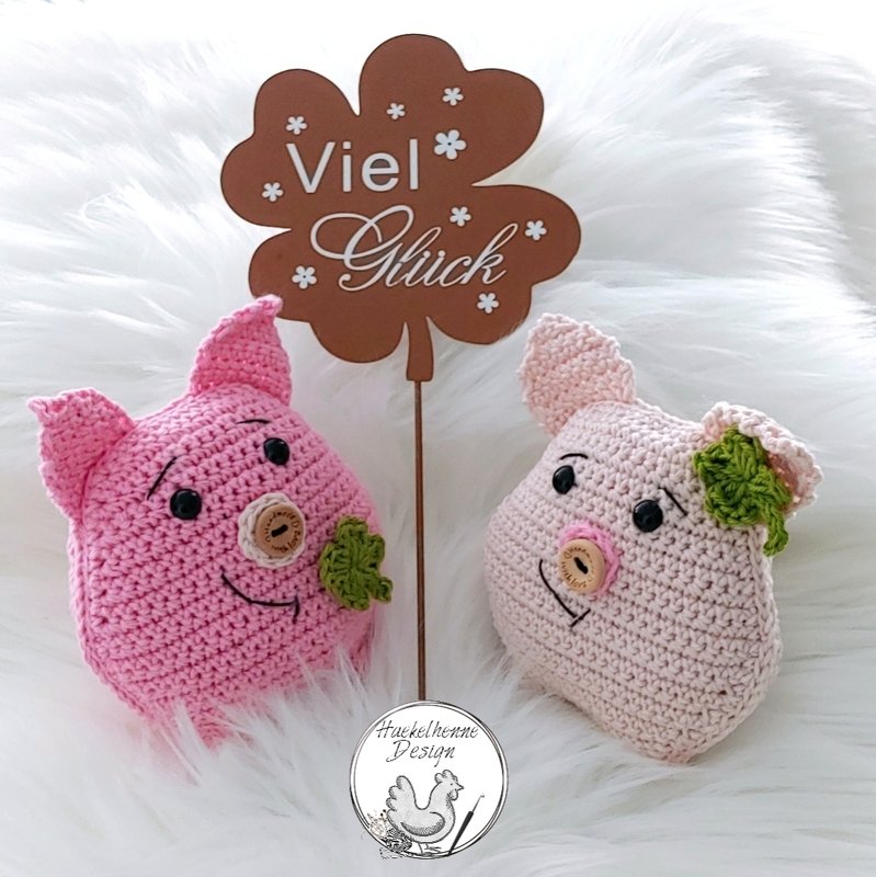 Zwei gehäkelte Amigurumi-Glücksschweinchen in Rosa mit Knopfnasen, Kleeblättern und 'Viel Glück'-Schild