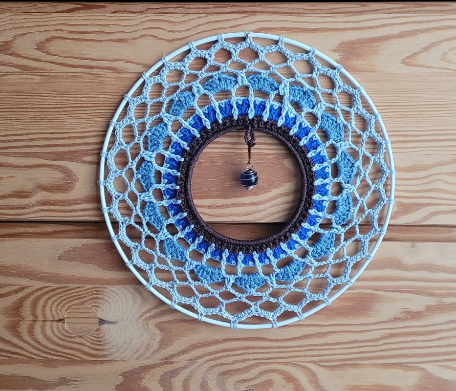 Handgehäkelte runde Boho-Wanddekoration in Blautönen mit zentraler dunkler Perle vor Holzuntergrund