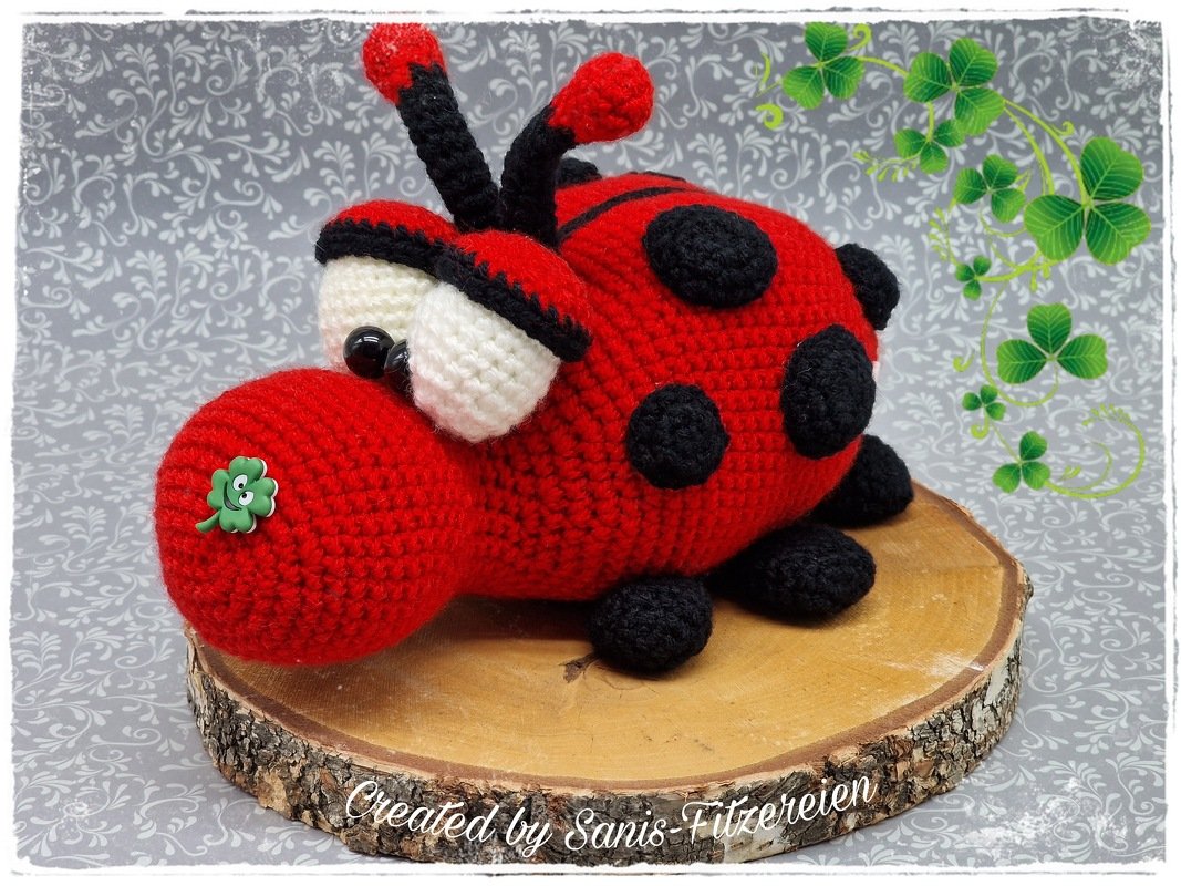 Roter gehäkelter Amigurumi-Marienkäfer mit schwarzen Punkten, weißen Augen und grünem Kleeblatt-Button auf einer Baumscheibe.