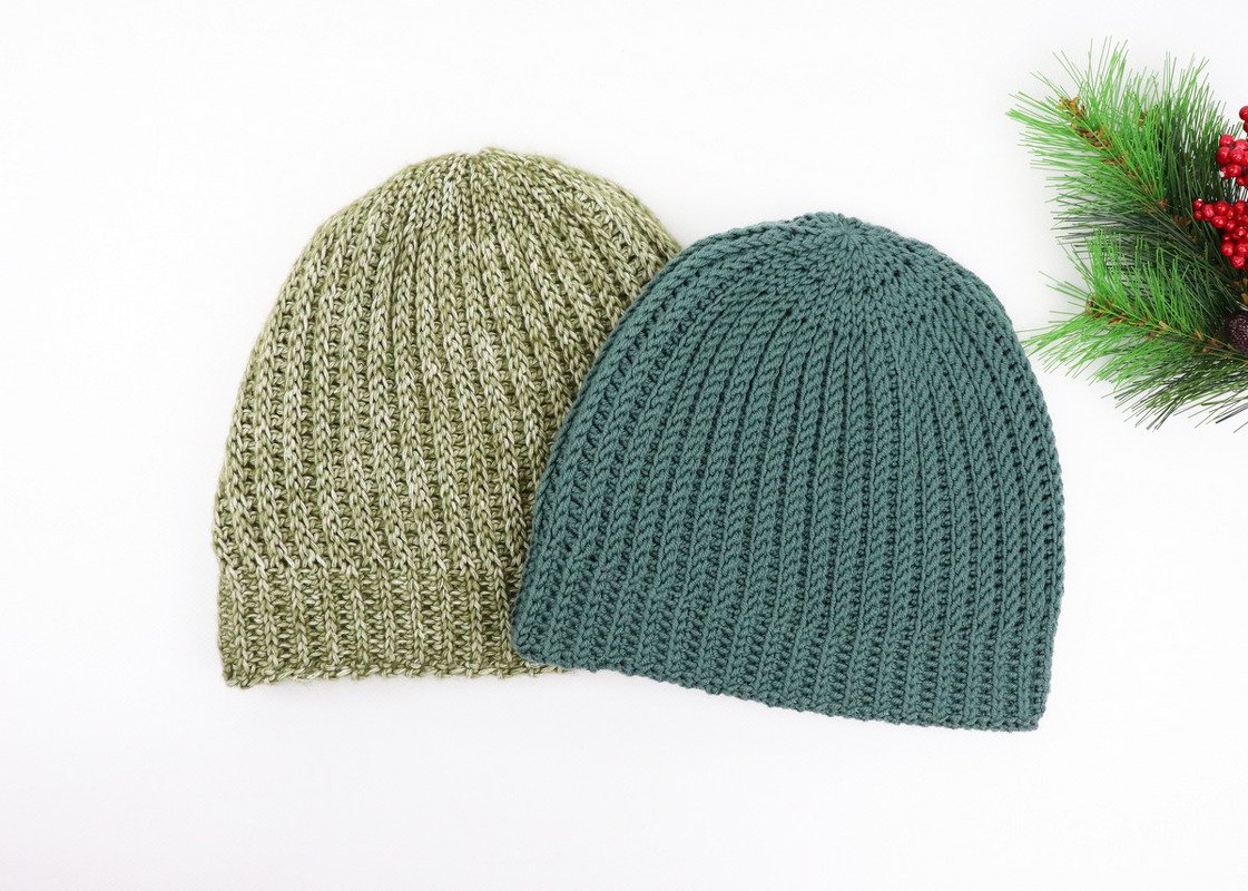 Two ribbed crochet beanies in teal and olive laid side by side on a white background with a sprig of evergreen and red berries