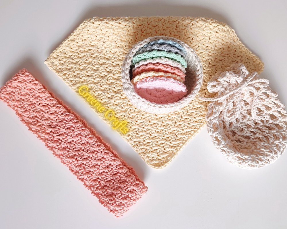 Pastel crocheted face pads stacked in a bowl with cream washcloth, mesh soap bag and coral textured strip on white background