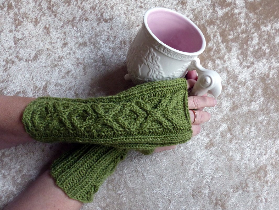 Gestrickte grüne, fingerlose Armstulpe mit Zopfmuster an einer Hand beim Halten einer weißen Tasse mit rosa Innenraum