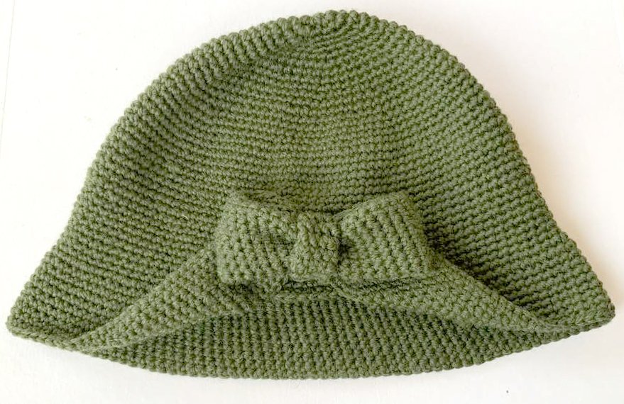 Eleganter gehäkelter Cloche-Hut in gedecktem Grün mit Schleife, auf hellem Hintergrund präsentiert.