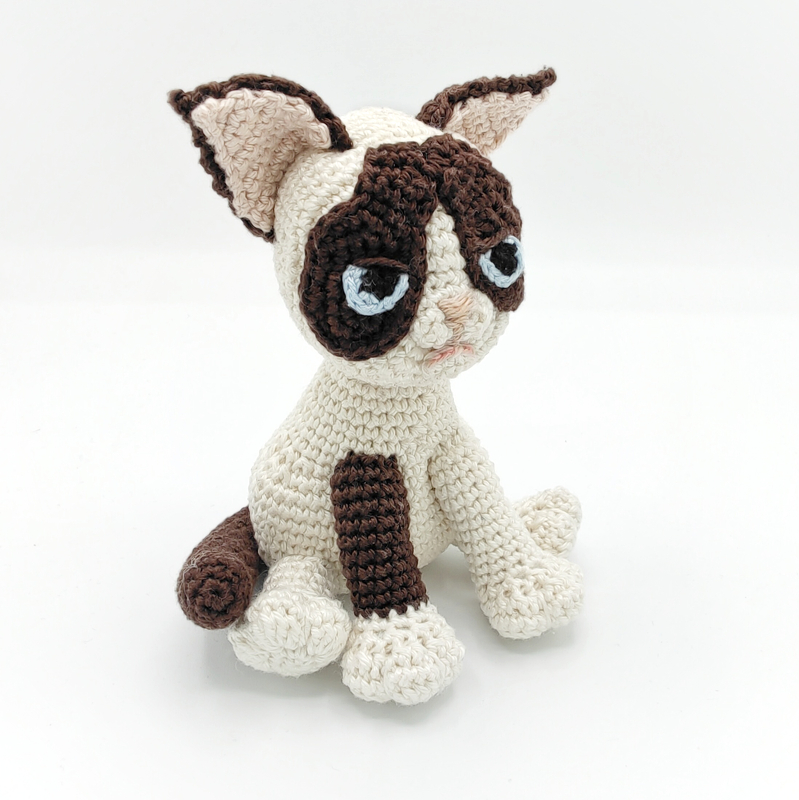 Sitting cream and brown amigurumi cat with pale blue stitched eyes on white background