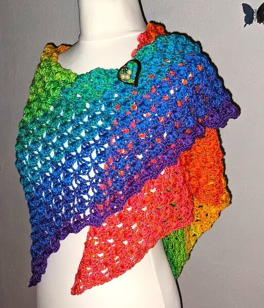 Gehäkeltes Dreieckstuch mit Regenbogen-Farbverlauf, auf weißer Schneiderpuppe drapiert und mit Herz‑Brosche befestigt