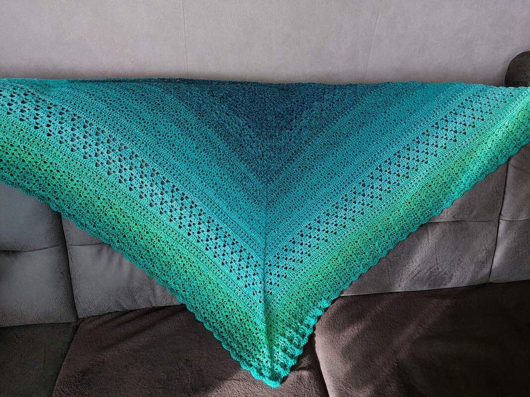 Gehäkeltes Dreieckstuch mit Türkis‑zu‑Grün‑Farbverlauf und Lochmuster auf einem Sofa drapiert.