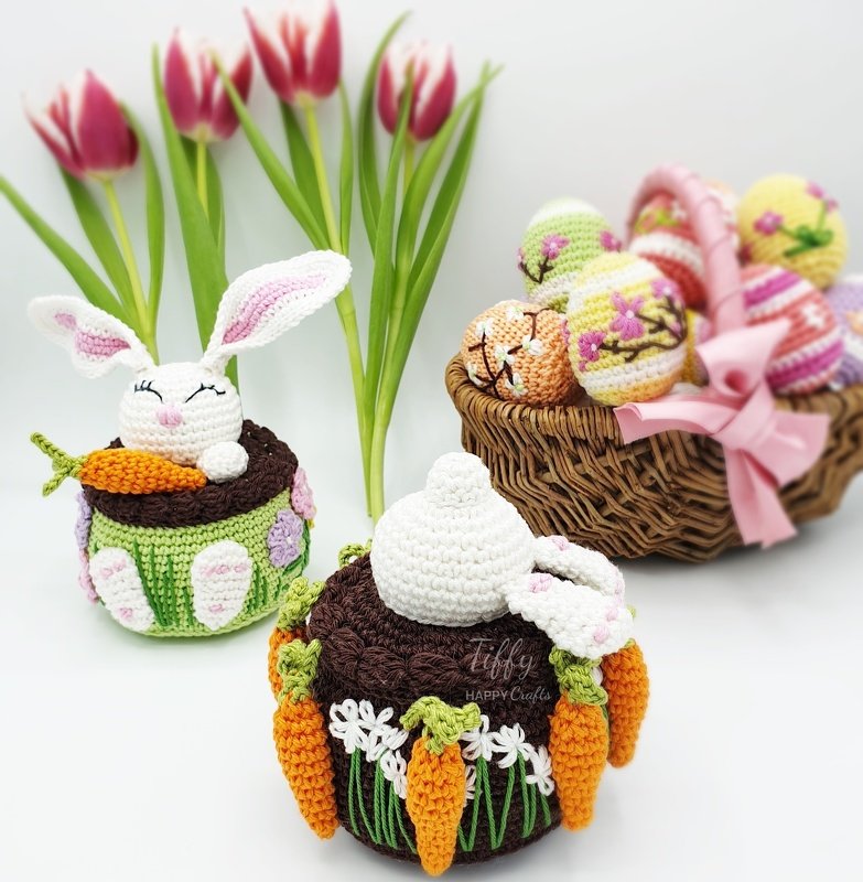 Two crocheted amigurumi rabbits, one peeking and one digging into a burrow, with carrot decorations and a basket of decorated eggs behind them