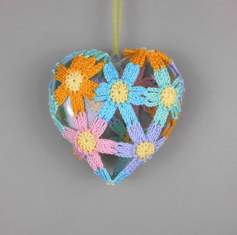 Transparenter Herzanhänger mit offener Hülle aus gehäkelten Blumen in Pastellfarben, hängend an einem gelben Band