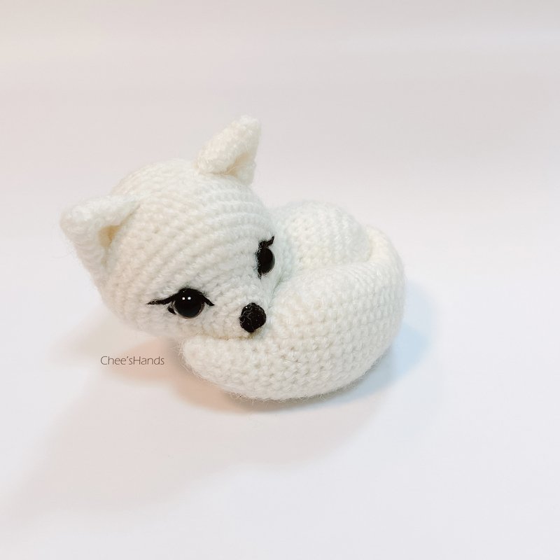 White crocheted fox amigurumi curled into a ring with black stitched eyes and nose