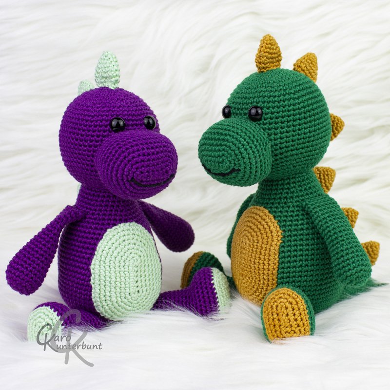 Zwei gehäkelte Amigurumi-Dinosaurier in Grün und Violett, sitzend auf weißem Hintergrund