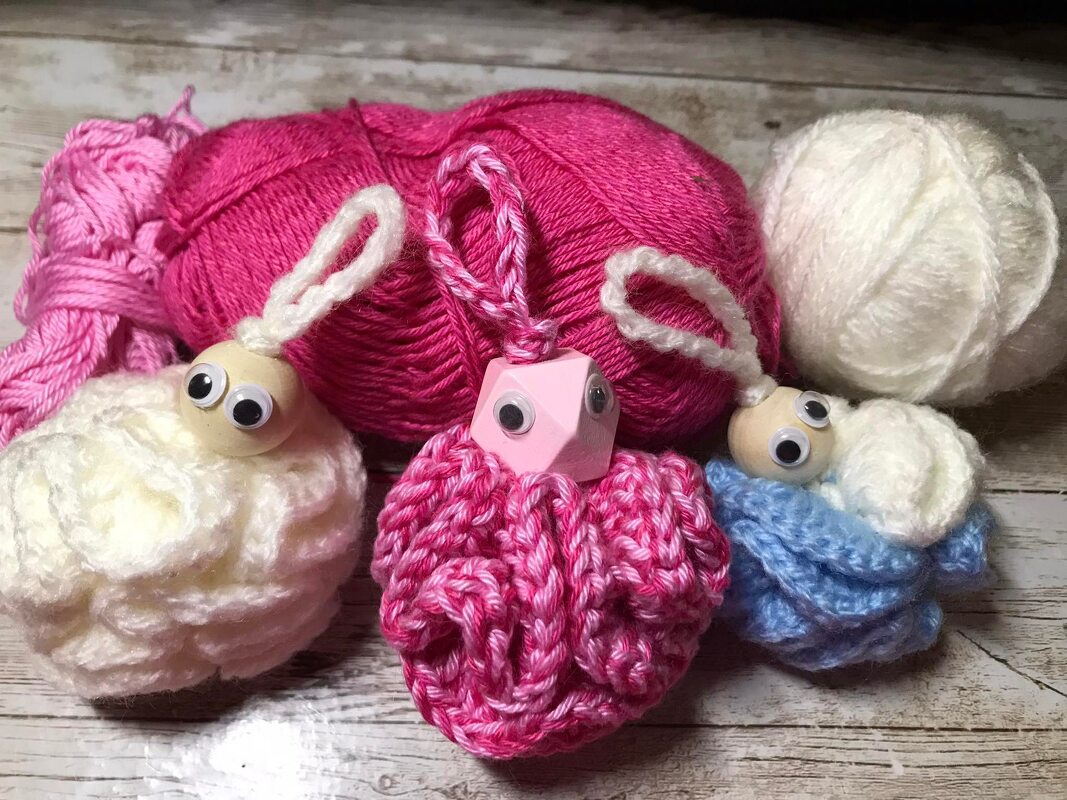 Drei gehäkelte Anhänger in Rosa, Weiß und Blau mit Wackelaugen und Aufhängeschlaufen vor Wollknäueln auf Holz.