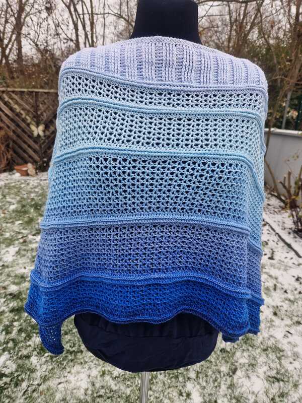 Gehäkelter Poncho in Blautönen mit Farbverlauf und variierenden Stichmustern auf einer Schneiderpuppe im Schnee.