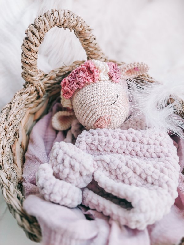Crocheted pink lamb snuggle blanket with a floral headband in a woven basket with white feathers