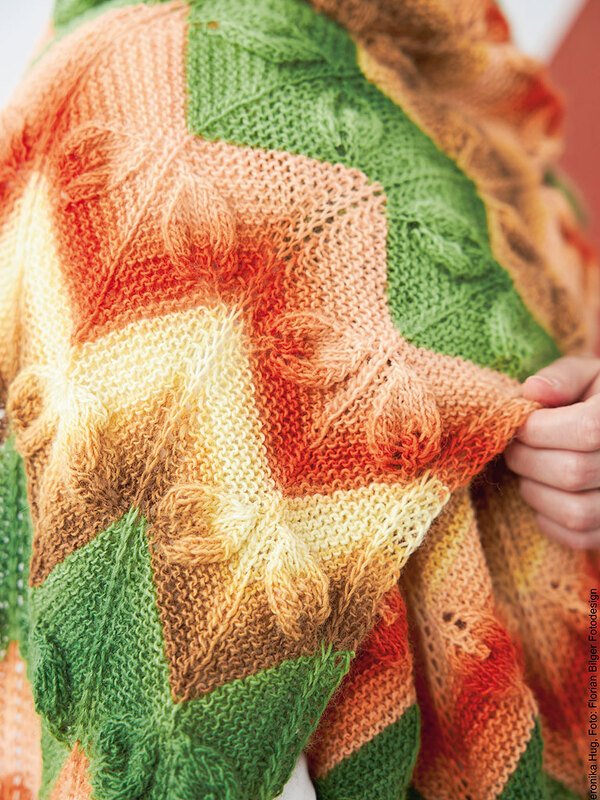 Nahaufnahme eines gestrickten Rechtecktuchs in Grün, Gelb, Orange und Braun mit Reliefmuster; eine Hand hält eine Ecke.