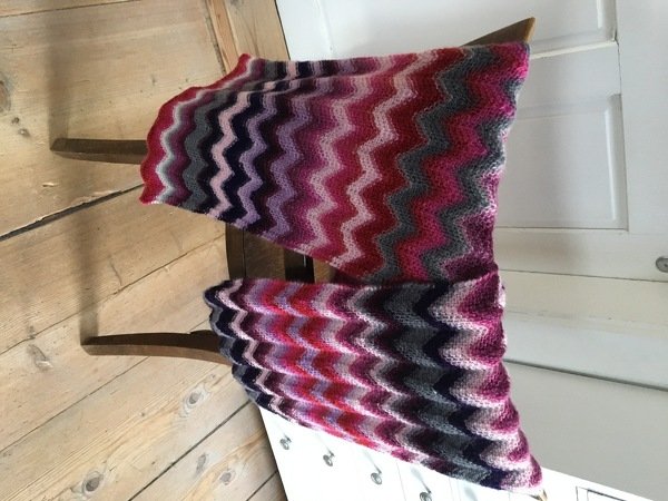 Mehrfarbiger gestrickter Loop‑Schal mit Zickzackstreifen in Rosa, Violett und Grau auf Stuhl drapiert