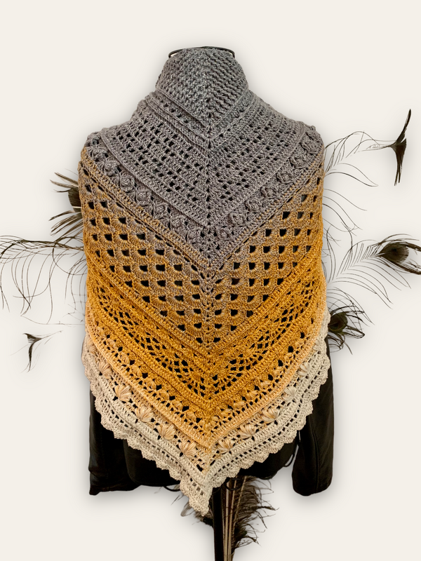 Triangle crocheted shawl on a mannequin with an ombre gradient from gray to golden yellow to cream, showing openwork stitch panels and a scalloped textured edge; feather accents around the display.