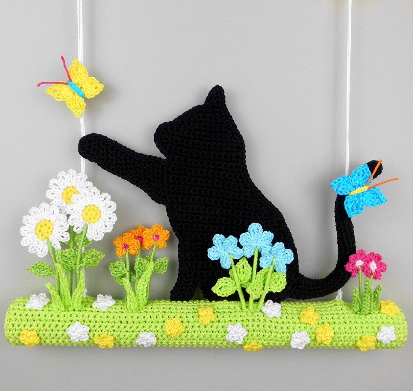 Handmade black crocheted cat silhouette reaching for a yellow butterfly, with a blue butterfly on its tail and colorful crocheted flowers on a green base, suspended by white cords.