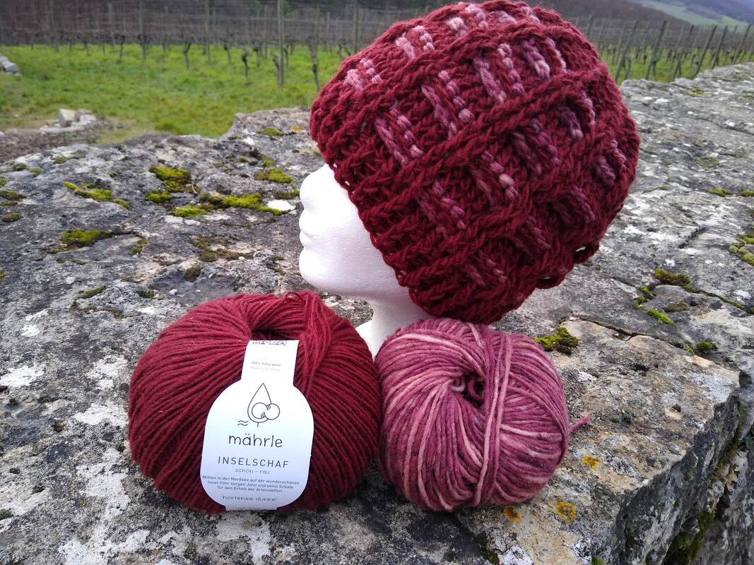 Gehäkelte burgundrote Mütze mit Rautenmuster auf weißem Schaumkopf, zwei Wollknäuel auf steinerner Mauer