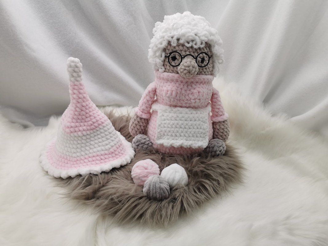 Gehäkelte Oma-Puppe mit rosa Kleidung, Brille, weißem lockigem Haar und weißer Schürze, sitzend neben einer rosa-weißen gestreiften Wichtelmütze und drei kleinen gehäkelten Kugeln auf flauschigem Untergrund.