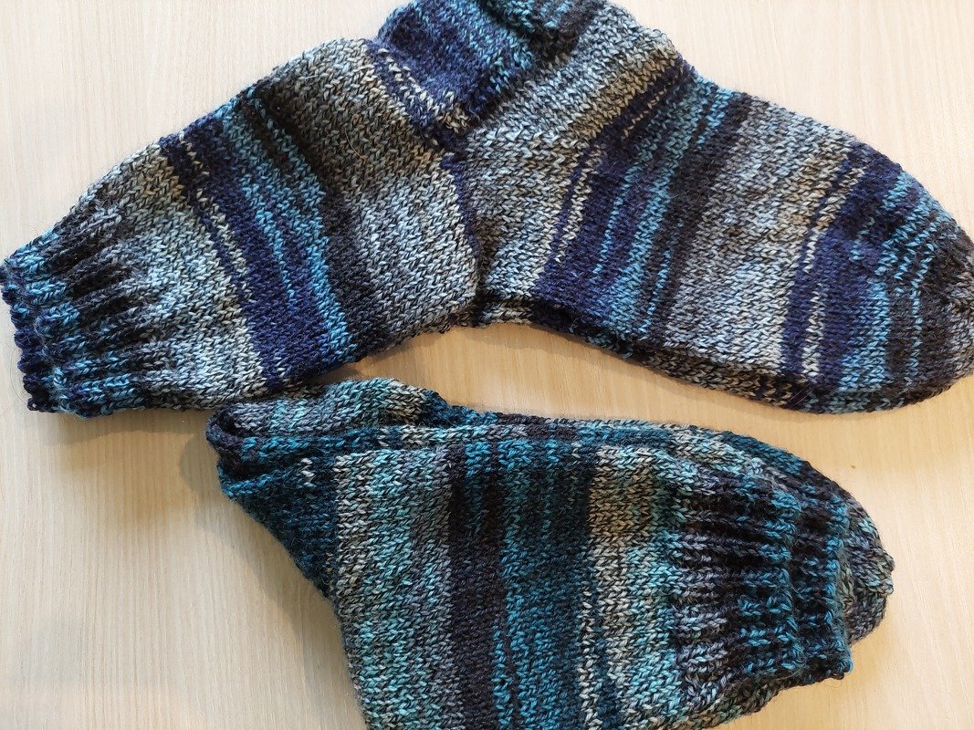 Zwei gestrickte Herrensocken in Blau-, Grau- und Schwarztönen mit Streifenmuster auf hellem Holzuntergrund