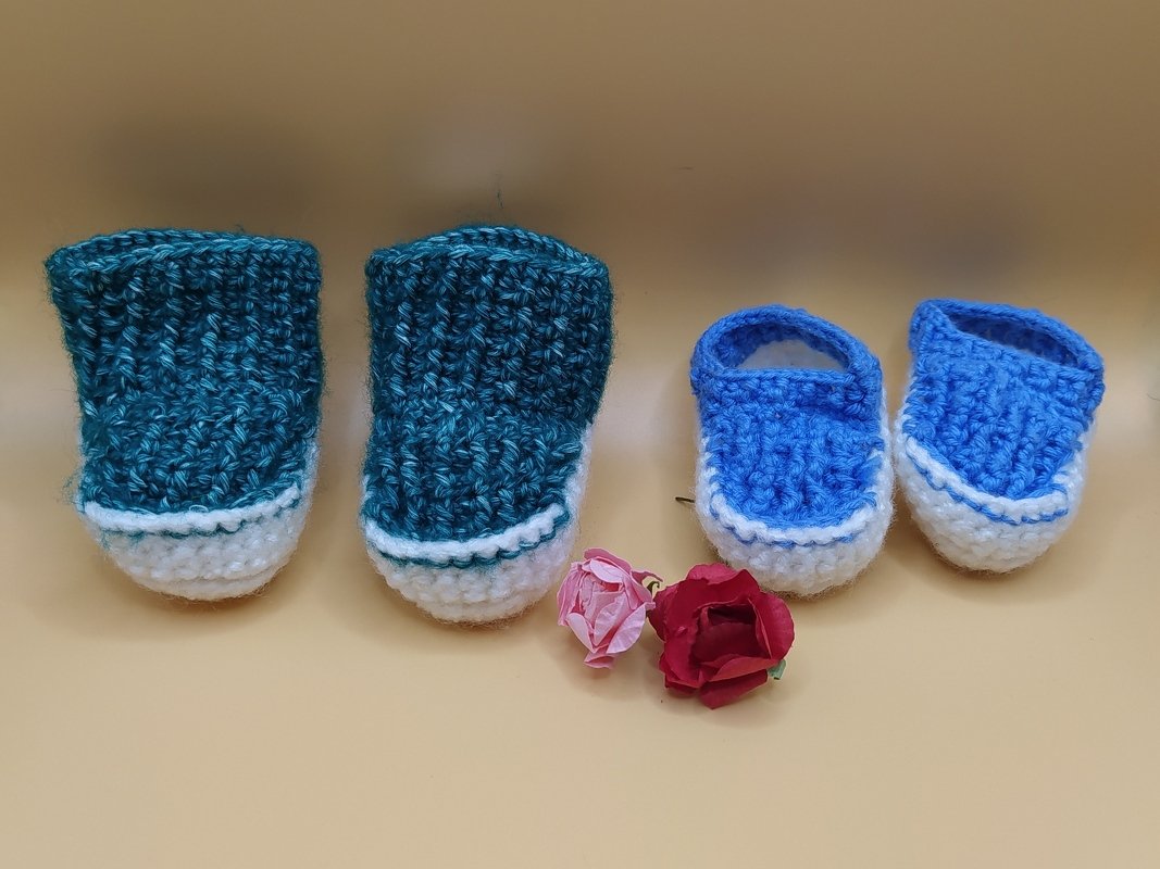 Zwei Paar gehäkelte Amigurumi-Puppenschuhe in Türkis und Blau auf beigem Hintergrund mit zwei Zierrosen.