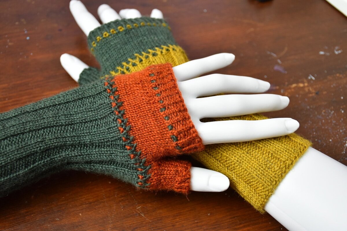 Zwei Paare gestrickter fingerloser Armstulpen in Grün, Orange und Senfgelb auf weißen Handmodellen