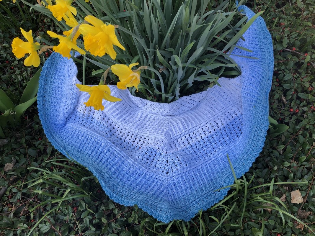 Gehäkeltes Dreieckstuch in Weiß und Blau mit Ajour- und Rippenmustern, umgeben von grünen Blättern und gelben Narzissen.