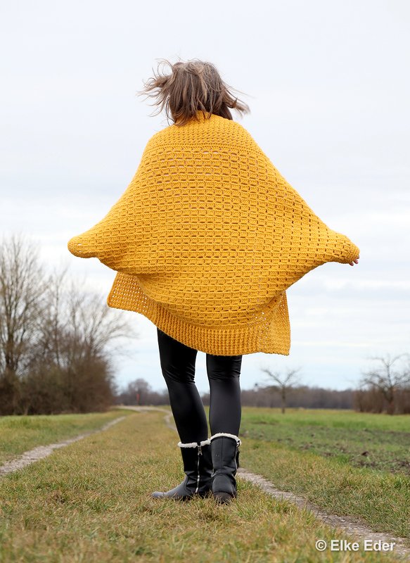 Rückansicht einer Person in weitem, gelben gehäkelten Seelenwärmer und schwarzen Leggings auf einem Feldweg.