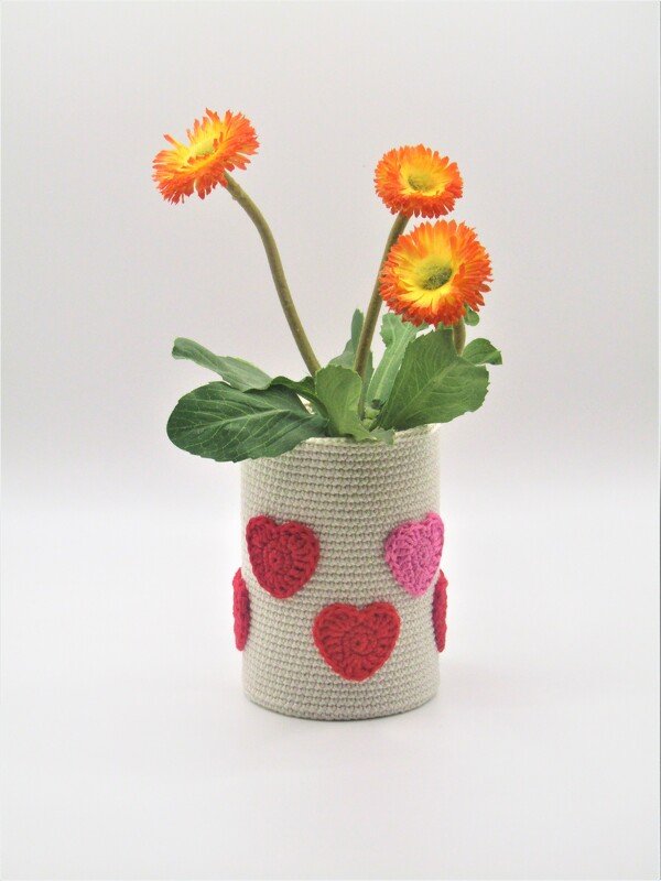 Cremefarbene gehäkelte Hülle in Zylinderform mit roten und rosa Herz-Applikationen, darin orange Blüten