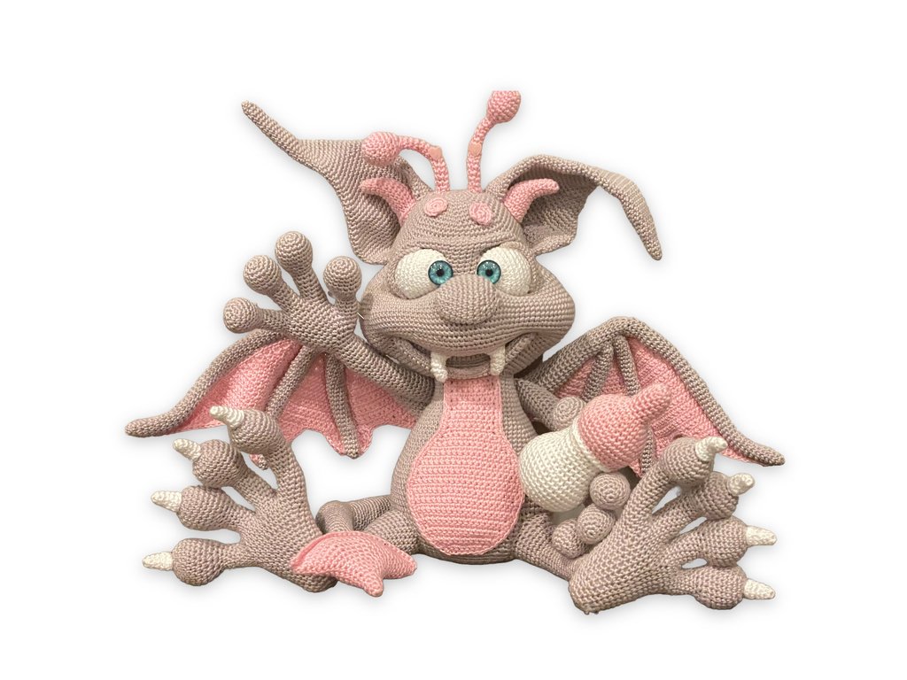 Gehäkelter Babydrache in Beige und Rosa, der ein Fläschchen hält.