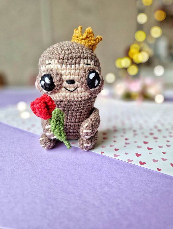 Crocheted sloth amigurumi wearing a crown and holding a red rose.