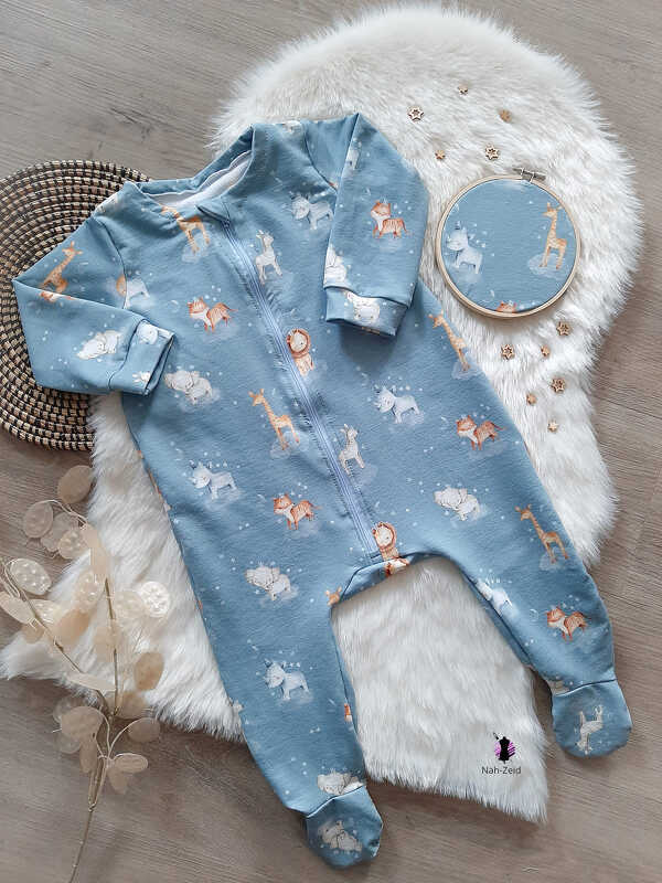 Hellblauer Baby-Strampler mit Safari-Tiermuster und durchgehendem Reißverschluss auf weißem Felluntergrund, daneben ein Stickrahmen mit passendem Stoffmuster.