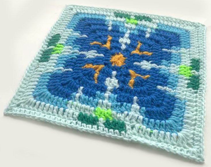 Gehäkeltes Quadrat in Blau, Gelb und Grün mit Sonnen- und Wellenmotiv auf hellem Hintergrund.