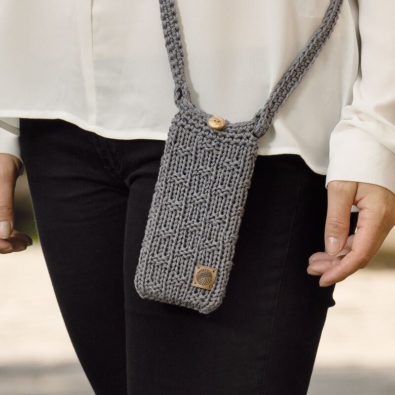 Grau gestrickte Handytasche mit Umhängeband und Holzknopf, getragen über einer weißen Bluse und schwarzen Hosen.