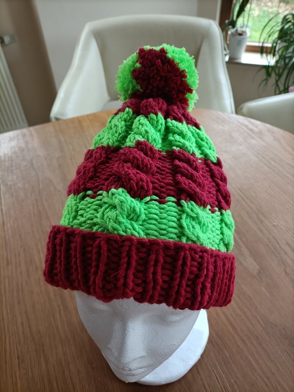 Gestrickte Kindermütze in Grün und Rot mit Zopfmuster und Bommel auf Mannequinkopf
