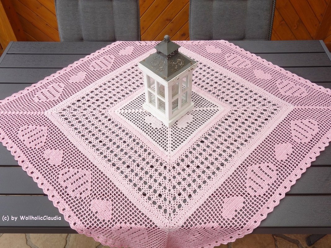 Rosafarbener gehäkelter Tischläufer mit Herz- und Filetmuster, diagonal auf einem dunklen Tisch mit weißer Laterne.
