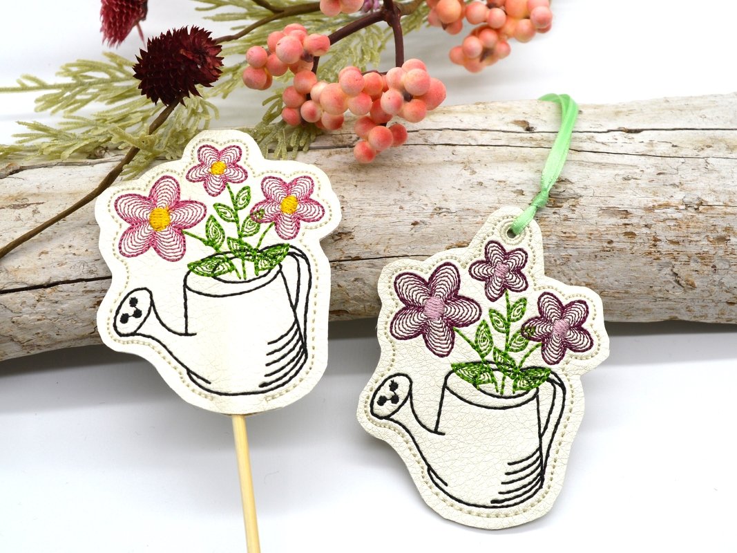 Zwei ITH-Gießkannen-Anhänger mit gestickten rosa Blüten, einer auf Holzstab, einer mit grünem Band, auf hellem Holz.