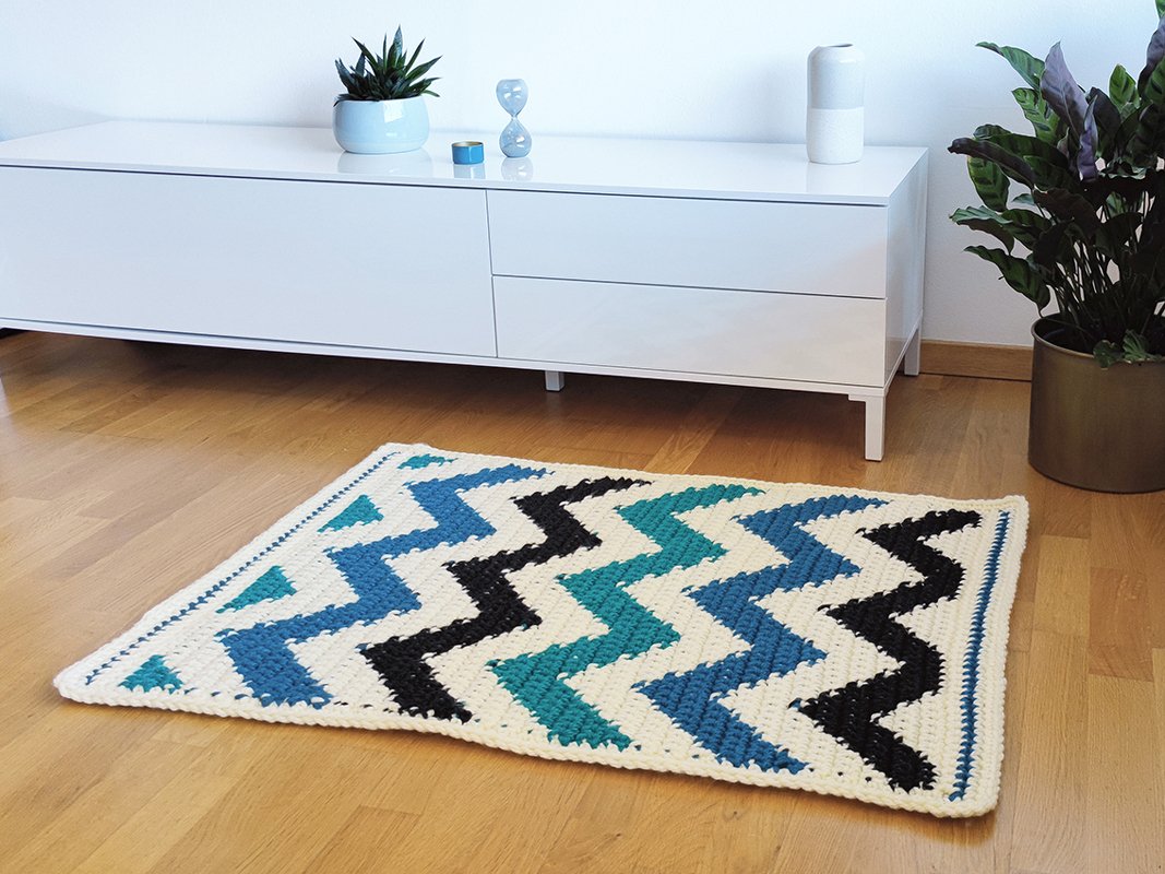 Gehäkelter Teppich in Creme, Blau, Türkis und Schwarz mit Zickzack-Muster auf Holzboden vor weißem Sideboard