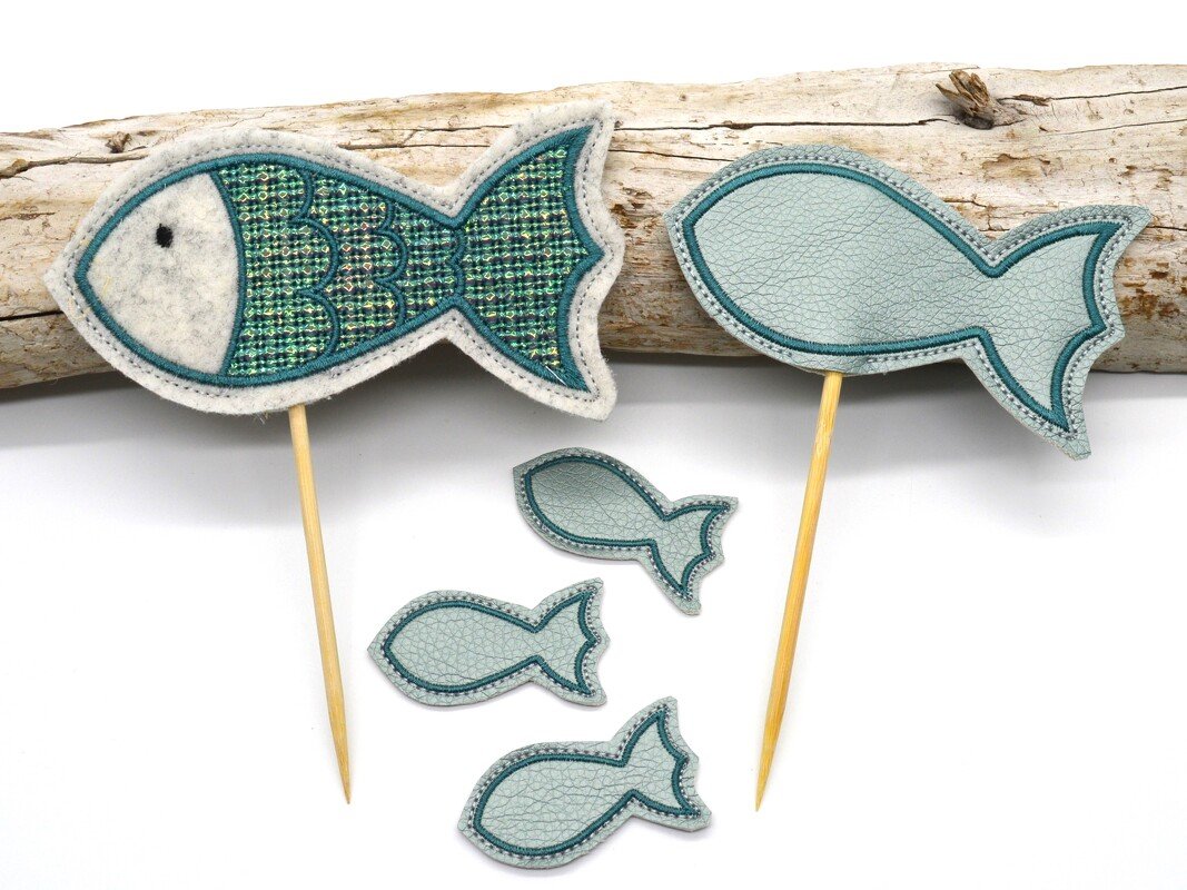 Zwei bestickte Fischfiguren auf Holzstäben und drei kleine Fischapplikationen in Türkis- und Hellblautönen vor einem Stück Treibholz auf weißem Hintergrund.
