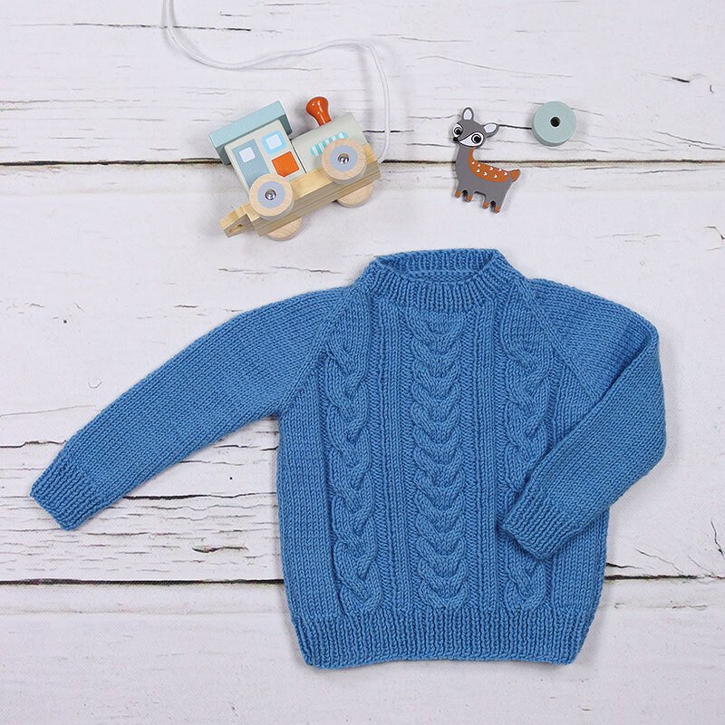 Blauer handgestrickter Kinderpullover mit Zopfmuster auf weißem Holzuntergrund, dekoriert mit Holzspielzeug