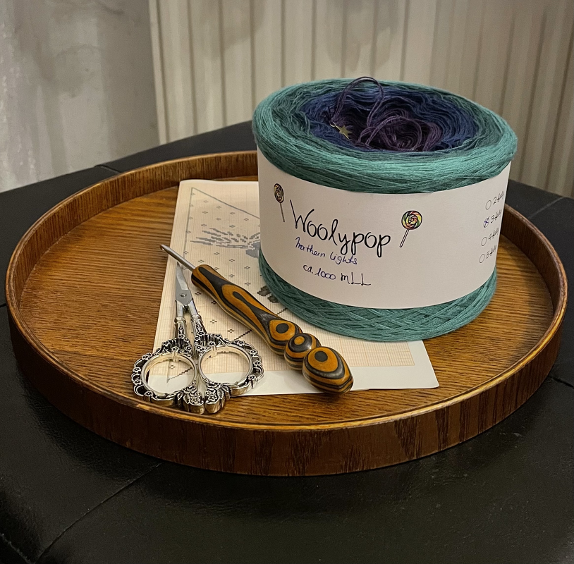 Rundes Holztablett mit Garnknäuel mit Farbverlauf, Holz‑Häkelnadel, verzierter Stickschere und Musterdiagramm