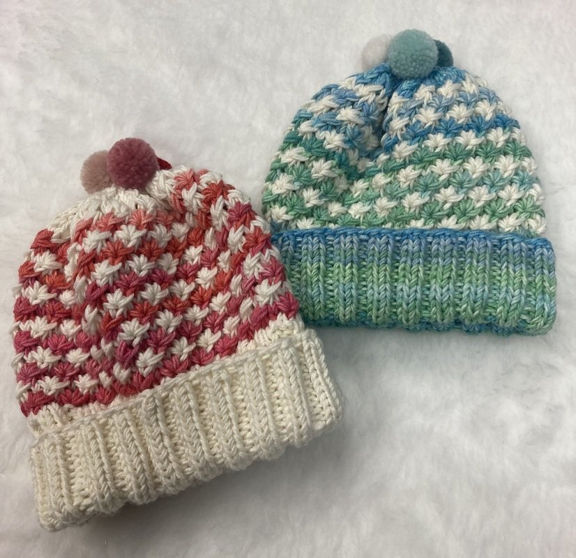 Zwei gestrickte Mützen in Rosa-Creme und Blau-Grün mit Pompons auf hellem, weichem Untergrund.